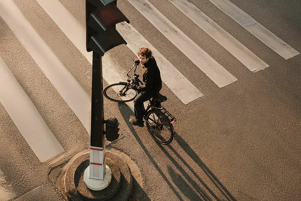 Aerial shot of a cyclist on the DUOTTS C29 Lite electric bike waiting at a city crosswalk during sunset, showcasing its sleek black frame, rear rack, and urban commuter design