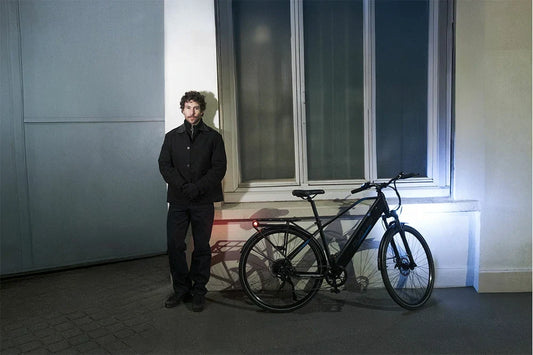 Curly haired man in black coat beside black DUOTTS urban commuter electric bicycle with rear rack, illuminated headlight and red tail light, dimly lit building wall with window, nighttime city scene